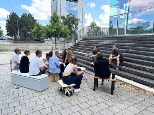 Gruppenfoto Menschen vor Hochschule sitzen auf Stühlen Carsten_Schelling_Entwurf 45_SoSe 2025