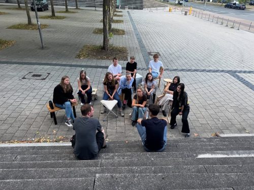 Gruppenfoto Menschen vor Hochschule sitzen auf Stühlen Carsten_Schelling_Entwurf 45_SoSe 2025